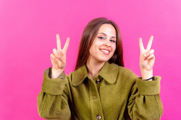 Giovane donna su sfondo rosa sorridente e mostrando segno di vittoria indossando giacca a vento verde