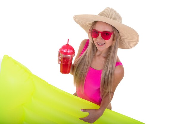 A young woman in a pink bathing suit and hat holds an air mattress and drinks lemonade