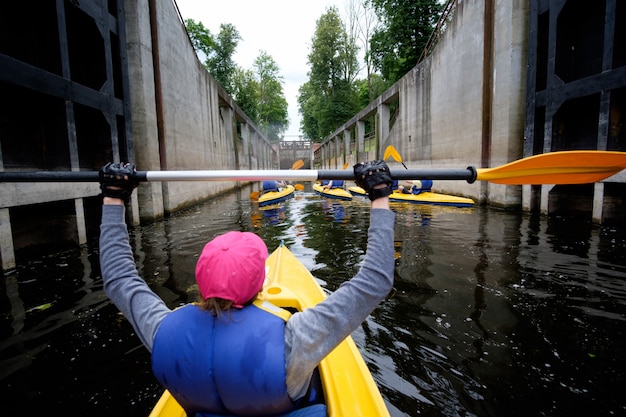 Young woman in pink cap rowing in kayak over the river channel in sluice, view from behind, kayak team