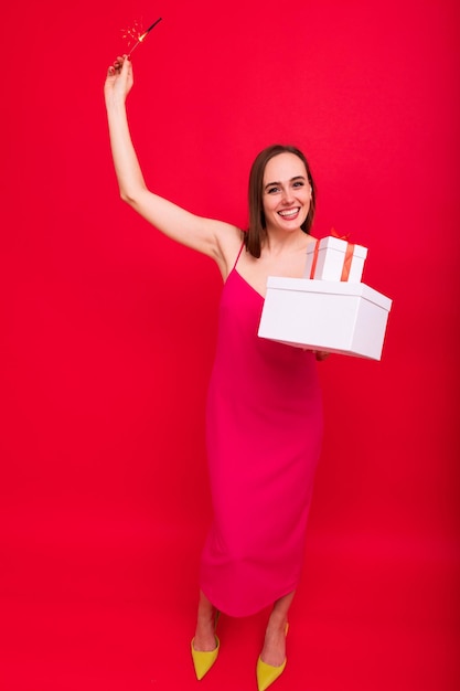 A young woman in a pink dress poses with Christmas gifts in boxes and sparklers on a red background