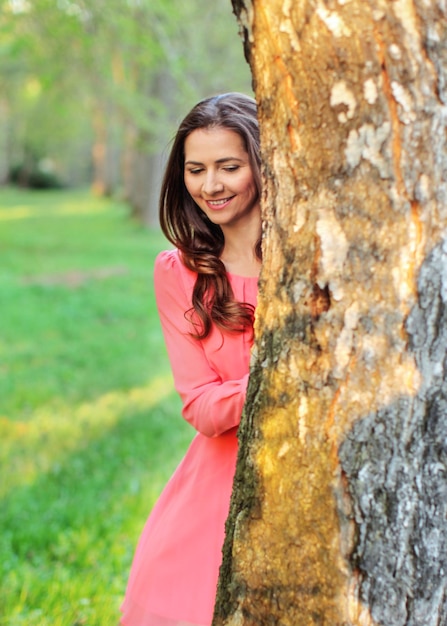 Young woman in pink dress, standing behind tree looking down, sunset light park in background.