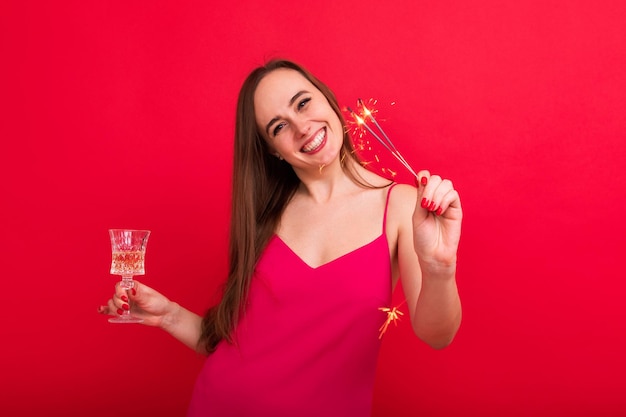A young woman in a pink dress with a glass of champagne and sparklers poses on a red background