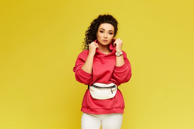 A young woman in a pink hoodie and white pants isolated at the yellow wall. Model girl with afro hairstyle in urban sportswear over yellow wall