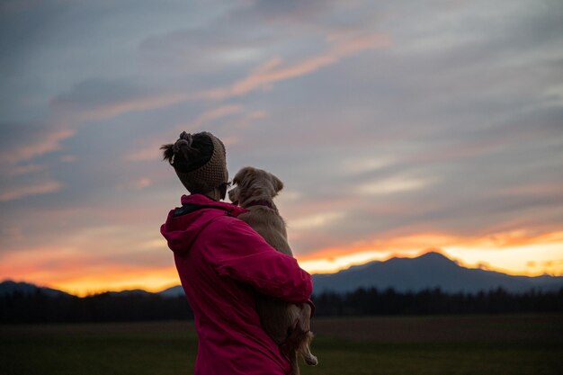 遠くを見ている美しいカラフルな夕方の空の下に立っている彼女の膝の上に彼女のかわいい犬を保持しているピンクのジャケットの若い女性