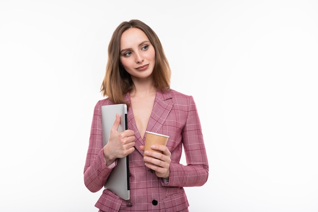 A young woman in a pink jacket holds a laptop and drinks coffee