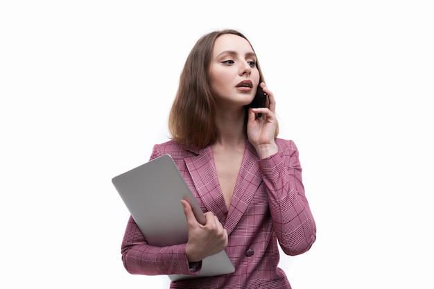 A young woman in a pink jacket is talking on the phone and holding a laptop on a white background Business style