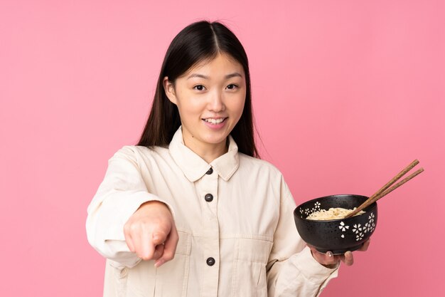 Giovane donna sul dito punti rosa con un'espressione sicura mentre si tiene una ciotola di noodles con le bacchette