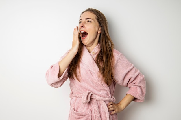 Young woman in a pink robe yawns on a white background