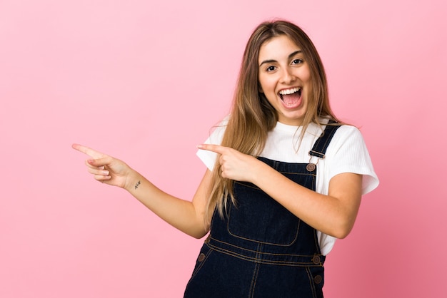 Young woman over pink wall surprised and pointing side