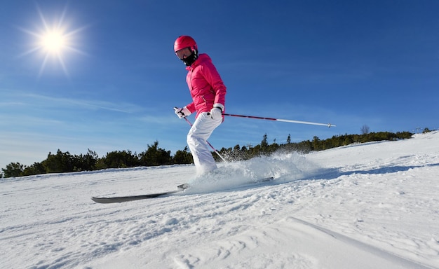 Young woman in pink winter jacket and helmet skiing snow spraying near, clear blue sky above