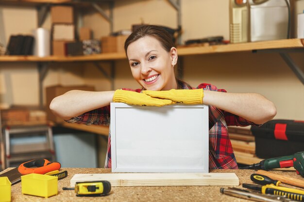 Молодая женщина в клетчатой рубашке, серой футболке, желтых перчатках, работающих в столярной мастерской на месте деревянного стола с пустой рамкой, различными инструментами. С пустым местом для текста. Скопируйте место для рекламы.