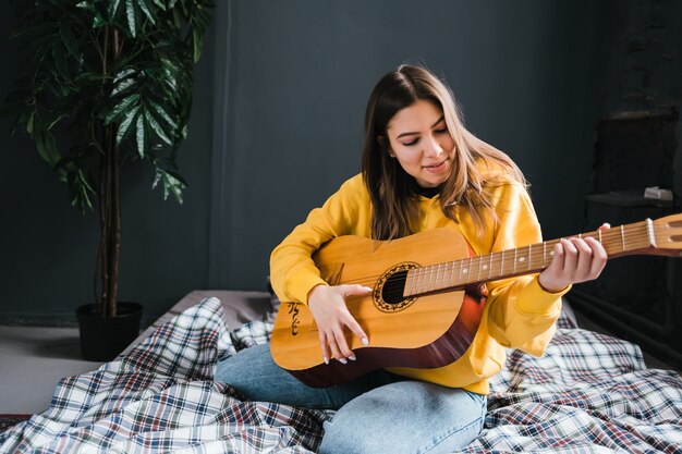 Giovane donna che suona la chitarra acustica a casa, seduta sul letto