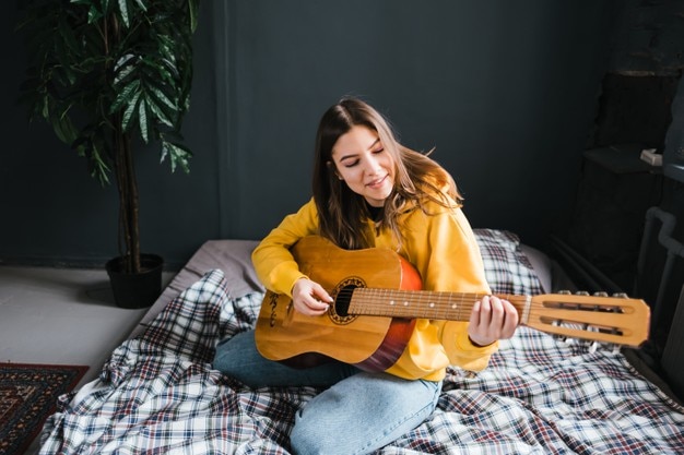 Young woman play on acoustic guitar at home, sitting on the bed