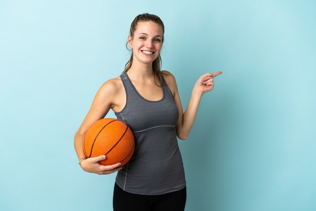 Young woman playing basketball isolated on blue pointing finger to the side