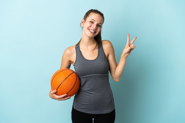 Молодая женщина, играющая в баскетбол, изолированная на синем, улыбается и показывает знак победы