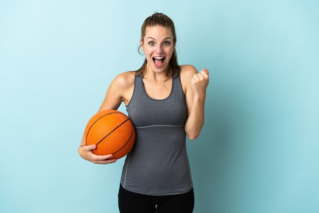 Young woman playing basketball isolated on blue wall celebrating a victory in winner position