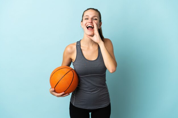 Young woman playing basketball isolated on blue wall shouting with mouth wide open