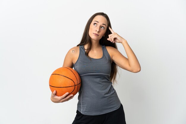 疑いと思考を持っている孤立した白い壁の上でバスケットボールをしている若い女性