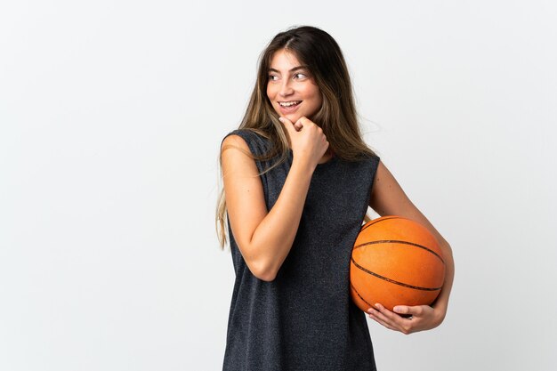 Молодая женщина, играющая в баскетбол, изолированная на белой стене, смотрит в сторону и улыбается