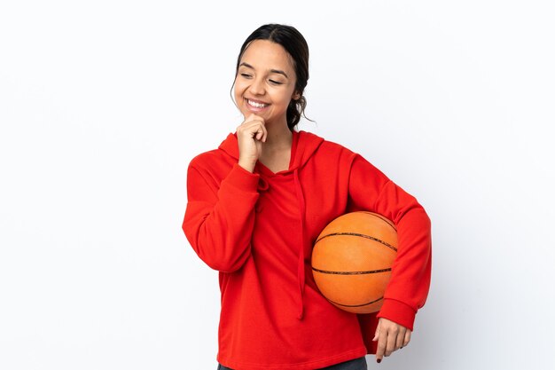 Young woman playing basketball over isolated white wall looking to the side and smiling