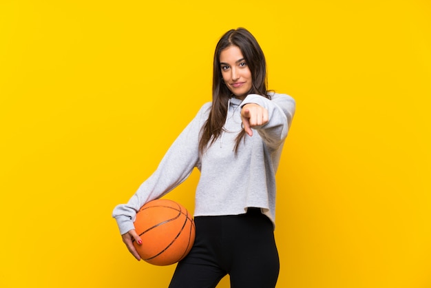 Young woman playing basketball over isolated yellow wall points finger at you with a confident expression