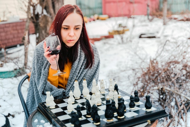 Young woman playing chess and drinking red wine in yard Female wrapped in grey plaid with alcohol sitting on street playing in board game in winter season