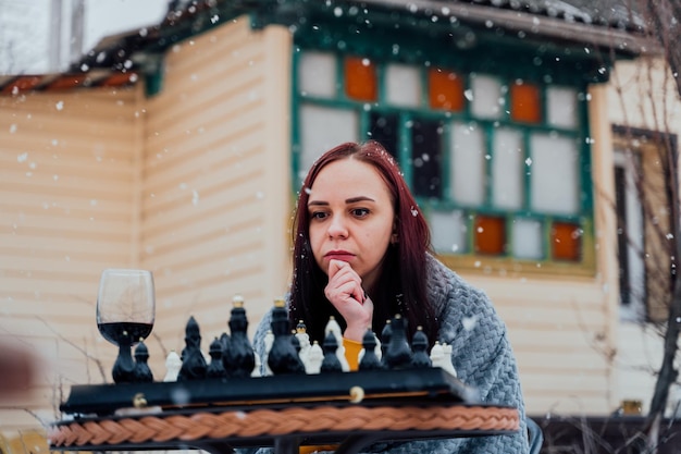 Young woman playing chess in yard Female wrapped in grey plaid sitting on street playing in board game in winter season