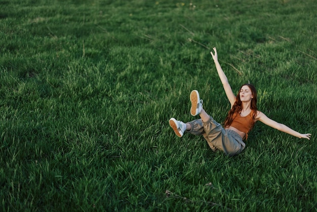 A young woman playing games in the park on the green grass spreading her arms and legs in different directions falling and smiling in the summer sunlight