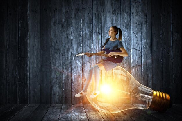 Photo young woman playing guitar sitting on light bulb. mixed media