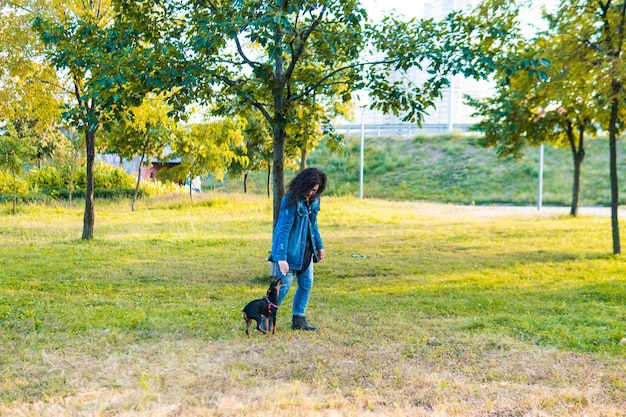 彼女の小さな犬を屋外で遊んで訓練している若い女性。公園で飼い主と黒と黄褐色のミニチュアピンシャー雌犬