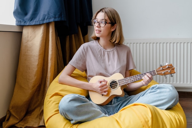 Young woman playing ukulele and staring at the window in the apartment