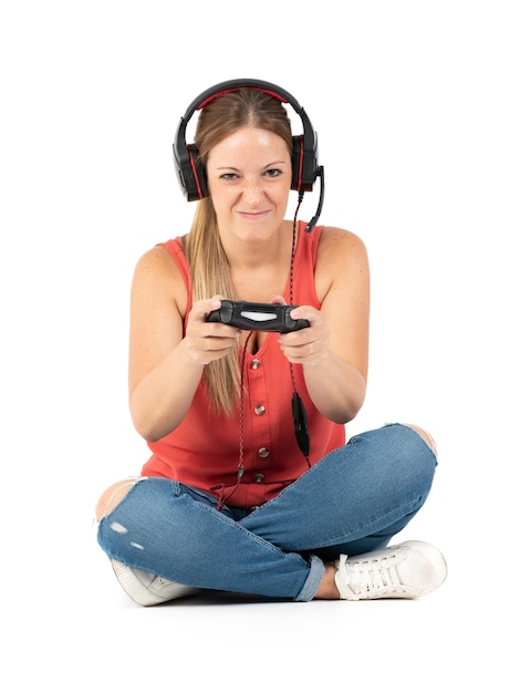 ジーンズの白い靴と赤いシャツと白い背景のコントローラーとヘッドフォンでビデオゲームをプレイする若い女性