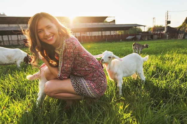 Young woman playing with goat kids on green spring meadow, smiling as animal chews her dress. Wide angle photo in strong sun backlight