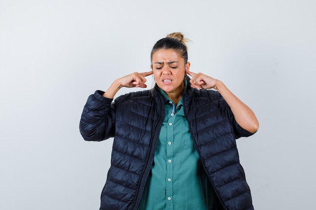 Young woman plugging ears with fingers in shirt, puffer jacket and looking annoyed , front view.