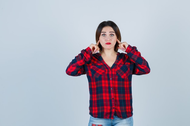 Giovane donna che tappa le orecchie con le dita indice in camicia a quadri, jeans e sembra tormentata. vista frontale.