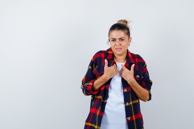 Young woman pointing at herself in checkered shirt and looking wistful , front view.