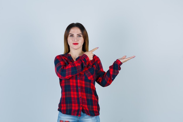 Young woman pointing right, spreading palm aside in checked shirt and looking sensible. front view.