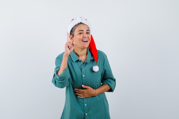 Young woman pointing up in shirt, Santa hat and looking joyful. front view.