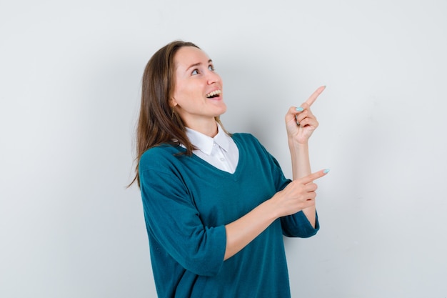 Photo young woman pointing at upper right corner, looking up in sweater over white shirt and looking happy. front view.