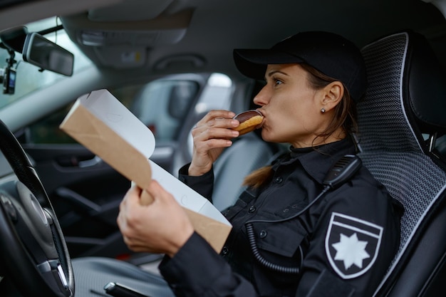 Agente di polizia della giovane donna che mangia la ciambella dolce che si siede in macchina durante la pausa pranzo sul lavoro