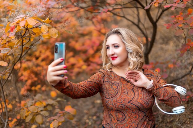 若い女性が秋の公園を背景にポーズをとって、スマートフォンで自分の写真を撮ります