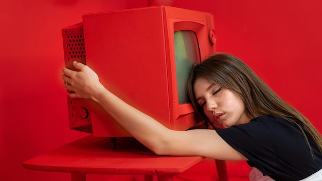 Молодая женщина позирует в студии с красными стенами и мебелью, держа в руках старый винтажный телевизор