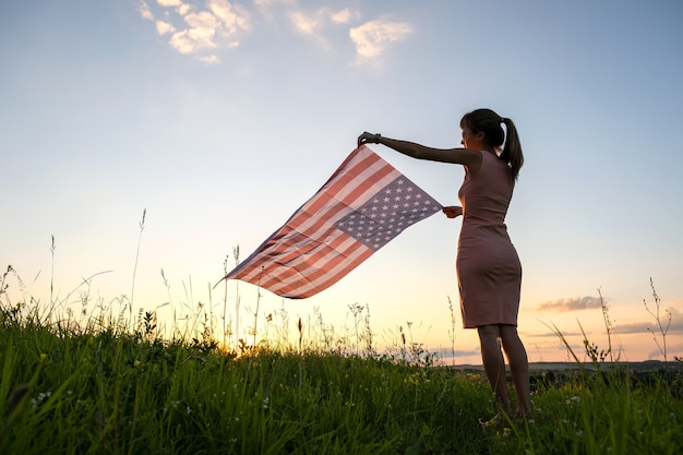 日没時に屋外でアメリカの国旗とポーズをとる若い女性。アメリカ合衆国の独立記念日を祝うポジティブな女性。