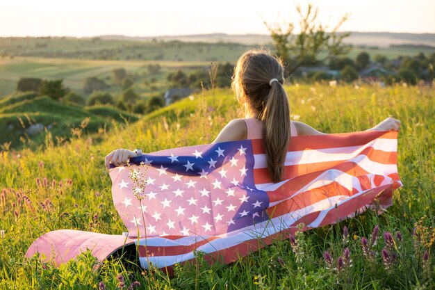 日没時に屋外でアメリカの国旗とポーズをとる若い女性。アメリカ合衆国の独立記念日を祝うポジティブな女の子。