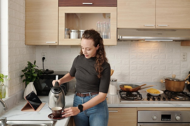 Young woman pouring herself cup of tea when cooking dinner at kitchen counter