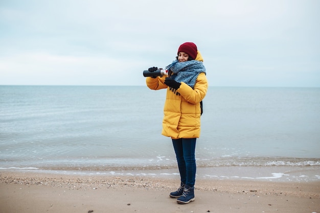 Young woman pours hot tea from a thermos into a cup and drinks on the seaside. Female tourist is having a wamn beverage at the winter cold beach. Hitch-hiking, travelling concept.