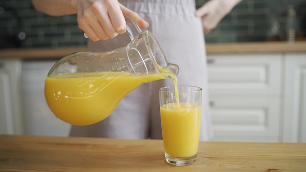 若い女性がデカンターからグラスにオレンジジュースを注ぎ、キッチンで飲みます。おはよう、幸せな週末、明るいキッチン。クローズアップ、HD