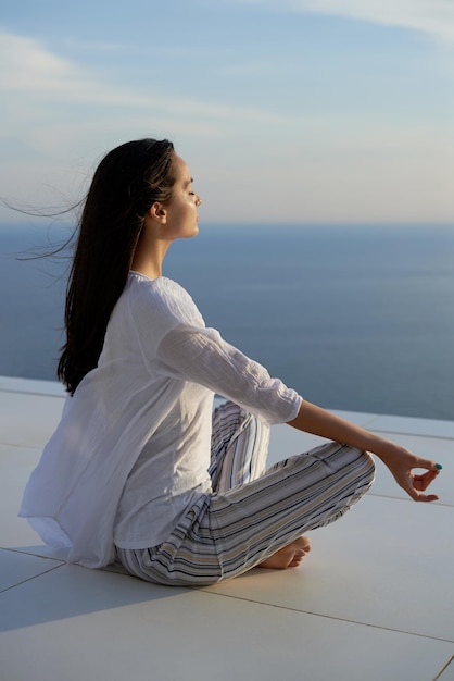 若い女性は、バック グラウンドで海の景色と夕日にヨガの瞑想を練習します。