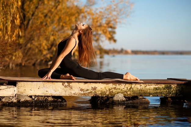 Photo young woman practicing yoga exercise at quiet pier in autumn park