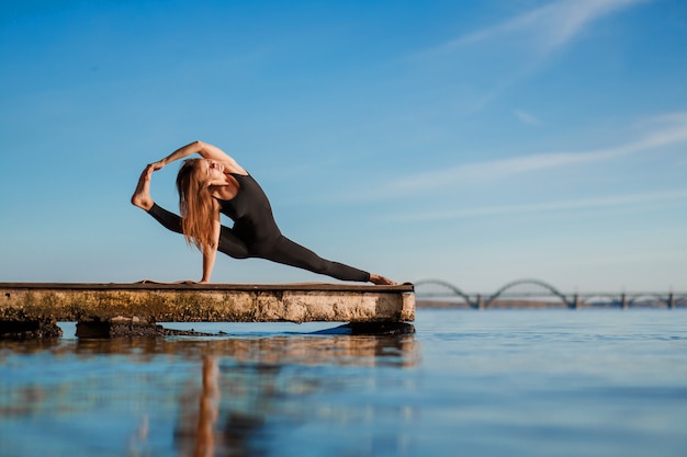 Esercitazione di yoga di pratica della giovane donna al molo di legno calmo con la città sport e ricreazione nella fretta della città